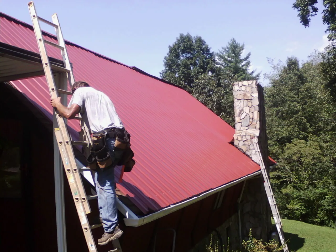 Roofer on a ladder beside a red metal roof and stone chimney — Soffit & Fascia Repair by Roofing Brothers in Valencia